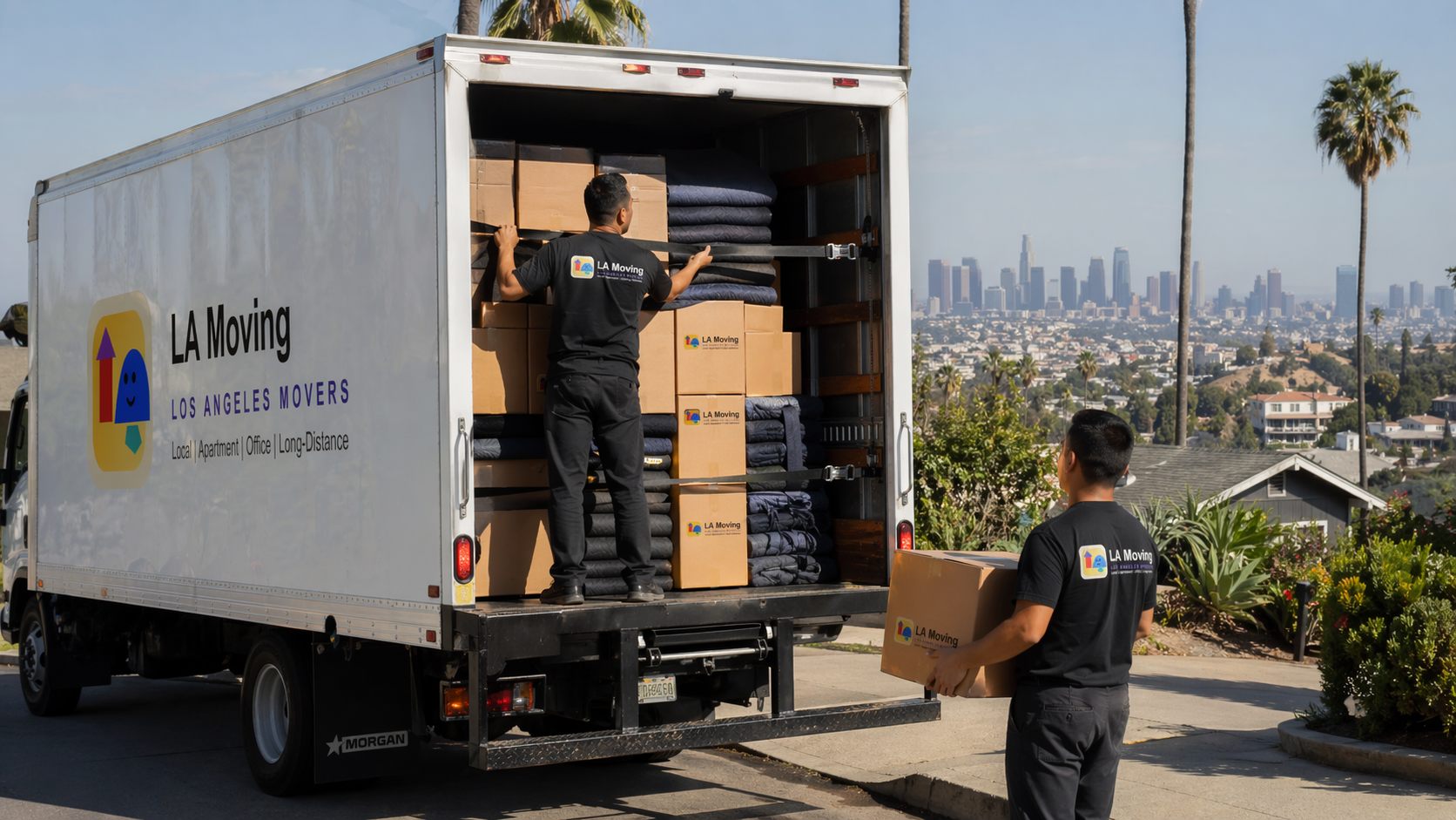 Two LA Moving movers in uniform loading branded boxes and moving blankets into a truck with the Los Angeles skyline behind them